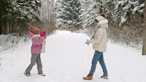 Larga vista lateral de la mamá y la hija asiática feliz con chaquetas hinchables, sombreros con pompones y bufandas sonriendo, divirtiéndose, jugando bolas de nieve en el bosque blanco de invierno - Imágenes, Vídeo