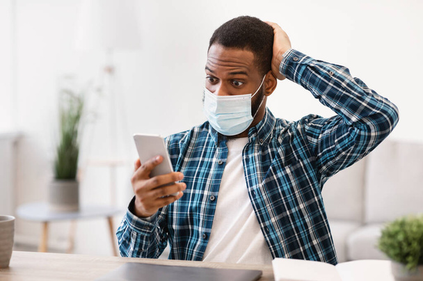 Czarny człowiek czyta wiadomości Coronavirus przez telefon nosząc maskę wewnątrz - Zdjęcie, obraz