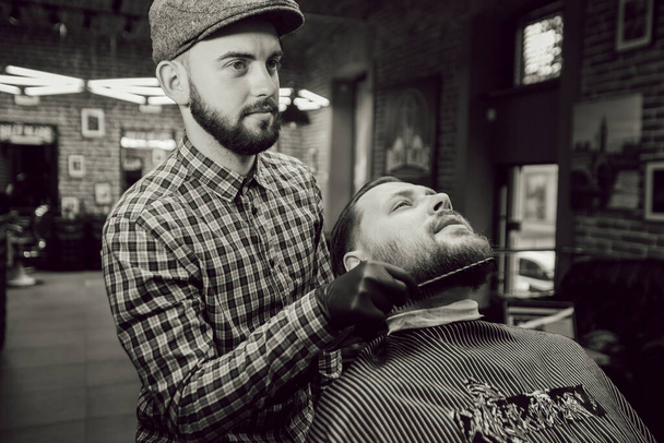 Kapsel in de kapperszaak. Close-up zicht van de jonge man met de baard, kapper kamt de klanten baard - Foto, afbeelding