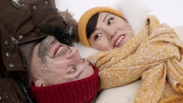 Alto ângulo médio close-up de feliz casal multiétnico vestindo roupas de inverno, deitado na neve, falando sorrindo - Filmagem, Vídeo