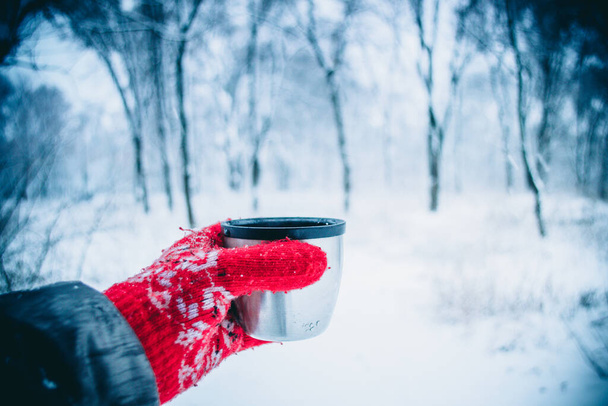 κορίτσι με μακριά μαλλιά σε κόκκινα γάντια με στολίδι κρατώντας έναν τουρίστα από ένα θερμός κύπελλο. Κατά την χιονόπτωση. Υπόβαθρο τουρισμού. Χειμερινό δάσος τοπίο θολή φόντο. Κρύο χειμερινό τοπίο - Φωτογραφία, εικόνα