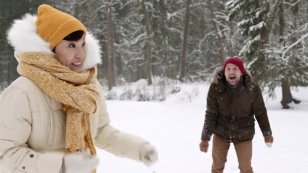 Mittlere Aufnahme einer fröhlichen Asiatin in Winterkleidung, die im Kiefernwald steht, lächelnd, kaukasischer Ehemann hinter ihr, der Schneeball auf Frau wirft, Paar spielt - Filmmaterial, Video