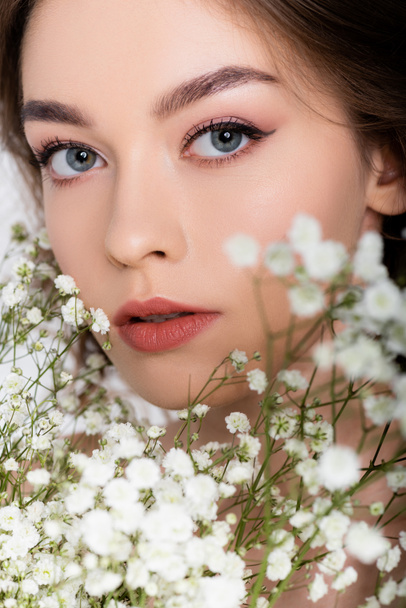 Młoda kobieta patrząc na aparat w pobliżu małych białych kwiatów na niewyraźnym pierwszym planie na białym tle  - Zdjęcie, obraz