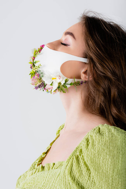 Young woman in medical mask with flowers standing with closed eyes isolated on grey  - Foto, Imagen