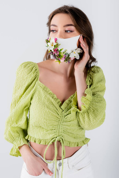 Young model with hand near face and flowers on medical mask isolated on grey  - 写真・画像