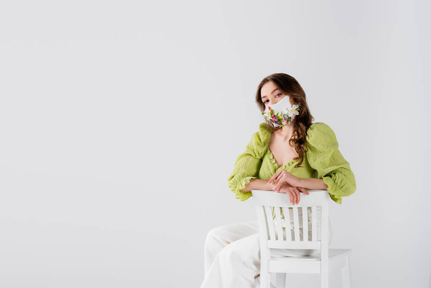 Brunette model with flowers on medical mask looking at camera on chair isolated on grey  - Фото, изображение