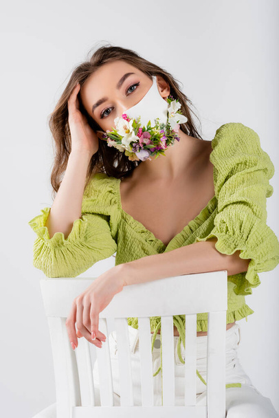 Stylish woman in medical mask with flowers posing on chair isolated on grey  - Foto, Imagem