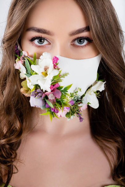 Egy nő portréja virágokkal orvosi maszkon, amint a szürkén izolált kamerát nézi  - Fotó, kép