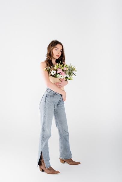 Volle Länge der stilvollen Modell mit Blumen in Bluse Blick auf Kamera auf grauem Hintergrund - Foto, Bild