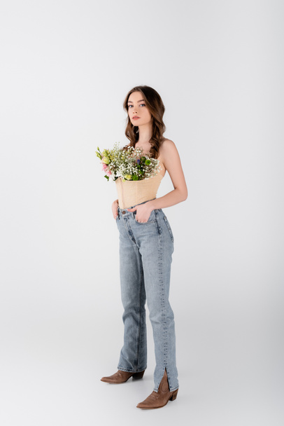 Молода модель з квітами в блузці і руками в кишенях джинсів дивиться на камеру на сірому фоні
 - Фото, зображення