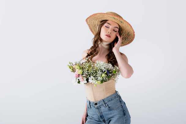 Modelka w dżinsach, czapce i kwiatach w bluzce pozowanie z zamkniętymi oczami na szarości  - Zdjęcie, obraz