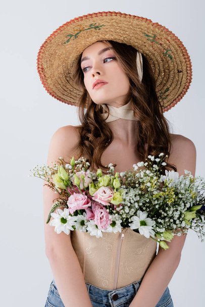 Modelo encaracolado em chapéu de sol com flores em blusa olhando para longe isolado em cinza  - Foto, Imagem