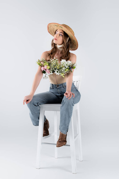 Young woman in sun hat and flowers in blouse looking away on chair on grey background  - Photo, Image