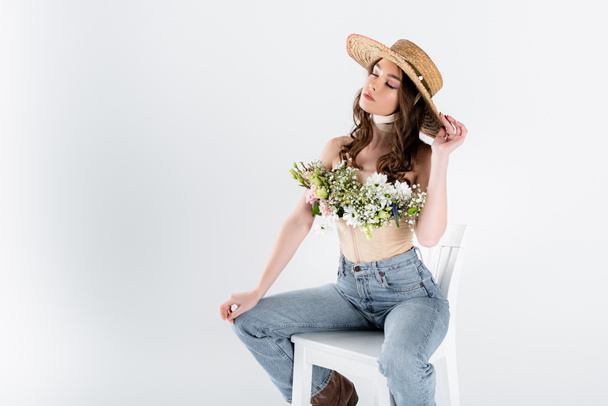 Young woman with flowers and sun hat sitting on white chair isolated on grey  - Photo, Image