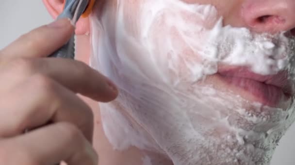 Een jongeman scheert zijn gezicht met een scheermes met schuim of scheergel in de badkamer. Gezichtsverzorging, scheren van gezichtshaar, stoppels, snor en baard. Ongeschoren jongen. - Video