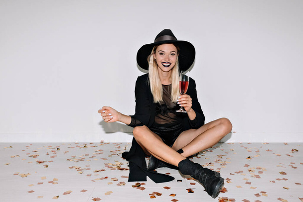Szczęśliwa uśmiechnięta dziewczyna z wakacyjnym makijażem i blond włosy noszące strój wieczorowy z kapeluszem siedzącym na podłodze z winem i konfetti, świętując wakacje  - Zdjęcie, obraz