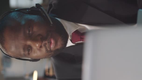 Вертикальний знімок афро-американського бізнесмена в бездротовому заголовку і офіційний костюм, виступаючи через відео дзвінок на ноутбук на роботі в офісі. - Кадри, відео