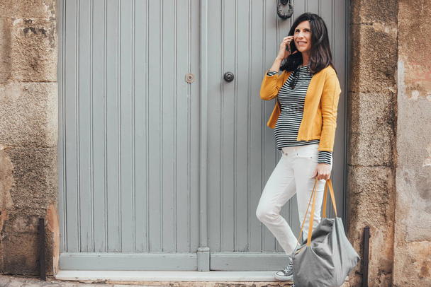 Young pregnant fashionable woman in stripes shirt, yellow jacket and bag, standing in front a door of an old town street, talking on the phone. Smiling brunet woman using mobile. Copy-space. - Fotó, kép
