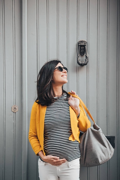 Young pregnant fashionable woman in sunglasses, stripes shirt, yellow jacket and bag, standing in front a door of an old town. Smiling brunet woman thoughtfully looking up the sky and holding her belly with one hand. - Foto, imagen