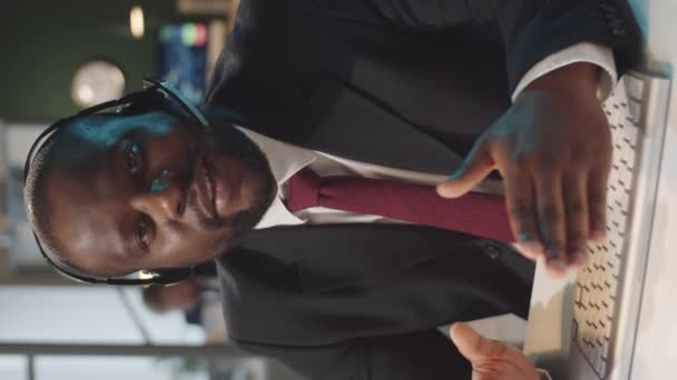 Вертикальный снимок черного бизнесмена в беспроводной гарнитуре и формальной одежде, разговаривающего на камеру во время веб-конференций в офисе - Кадры, видео