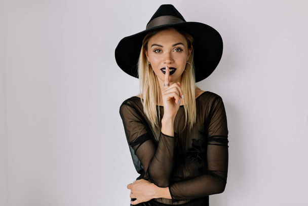 Spectacular cute girl wearing hat and black blouse holding finger near her lips and smiling over isolated background. High quality photo - Photo, Image