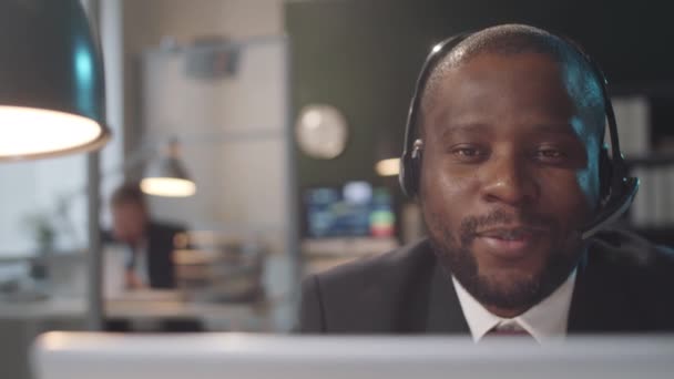 Tracking shot of Afro-American businessman in wireless headset and formalwear sitting at office desk and talking via online video call on laptop - Imágenes, Vídeo