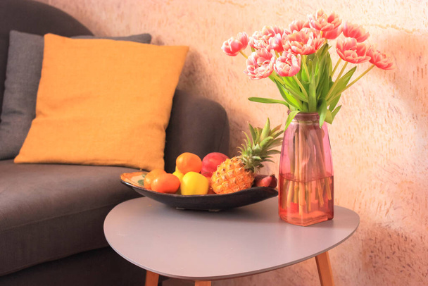 居心地の良いリビングルームのインテリア、グレーのソファと黄色の枕。ガラスの花瓶にピンクのチューリップの花束と異なるトロピカルフルーツを持つプレート-パイナップル、リンゴ、柑橘類-小さな灰色のテーブルの上. - 写真・画像