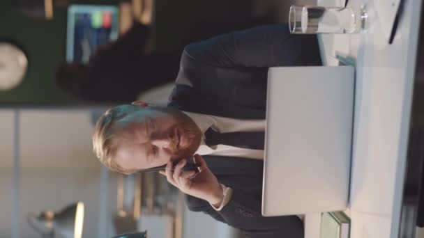 Plan vertical d'un homme d'affaires caucasien joyeux en tenue de cérémonie assis au bureau, parlant sur un téléphone portable et utilisant un ordinateur portable - Séquence, vidéo