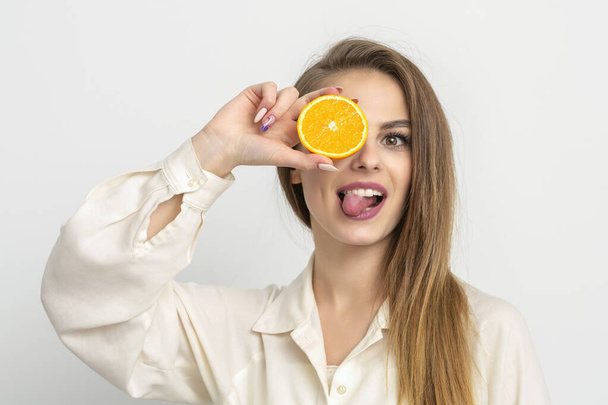 明るい白人の若い女性がオレンジのスライスで目を覆い、白い背景に白いシャツを着て舌を出します - 写真・画像