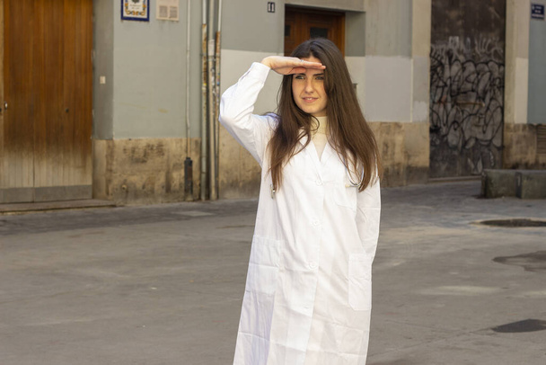 A shallow focus of a brunette female wearing a medical robe and shading her eyes with her hand outdoors - Fotó, kép