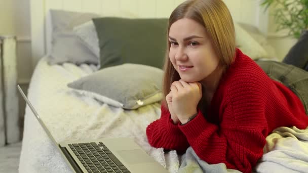 Jonge vrouw werkt op een computer, zittend op een bed, van een afstand werkend. Een meisje met lang haar in een rode trui en jeans werkt thuis. - Video