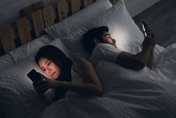 Konflikt zwischen jungen Paaren im Bett. Glücklich lächelnde Frau kehrte dem Mann den Rücken zu, las Nachricht auf Handy und versuchte, auf Bildschirm zu blicken. Betrug und Untreue. - Foto, Bild
