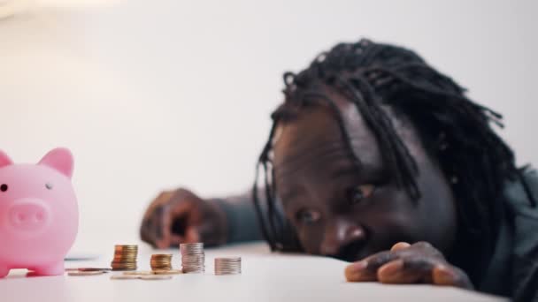 Zdesperowany czarny mężczyzna wygląda smutno w monetach w pobliżu skarbonki - Materiał filmowy, wideo