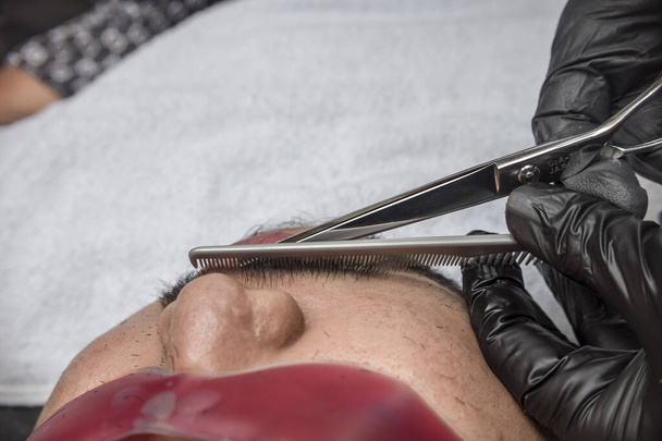Un peluquero recorta y moldea cuidadosamente el bigote de un hombre asiático. Peinado facial profesionalmente hecho en una peluquería o salón. - Foto, Imagen