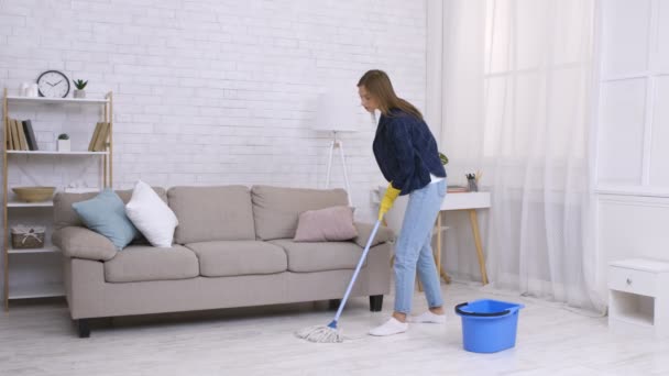 Nettoyage de printemps. Jeune femme au foyer nettoyage sol à la maison avec balai et seau, portant des gants en caoutchouc - Séquence, vidéo