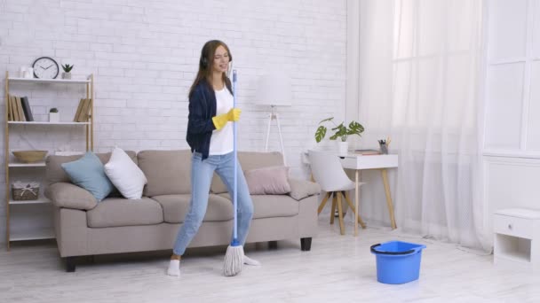 Le nettoyage printanier est amusant. Joyeux jeune femme dansant et chantant dans la serpillière comme micro, écoutant de la musique dans les écouteurs - Séquence, vidéo