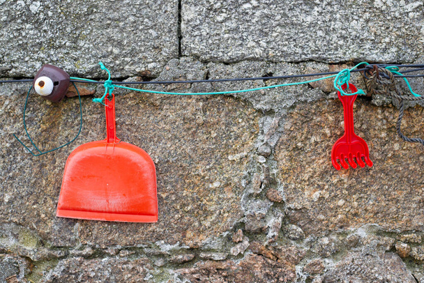 An orange plastic hand dustpan and a red plastic rake hanging from a green cord with a stone wall in the background - Photo, Image