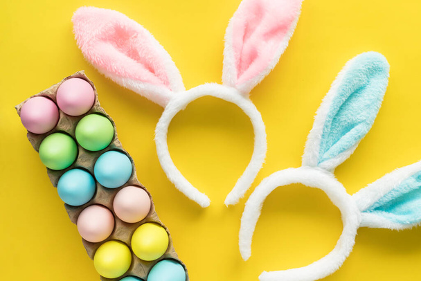 Colored Easter eggs and bunny ears headbands for children. Easter holiday flat lay on yellow background. Egg hunting concept - Photo, Image