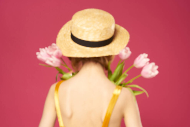 Pretty woman in yellow dress hat bouquet of flowers pink background back view - Photo, Image