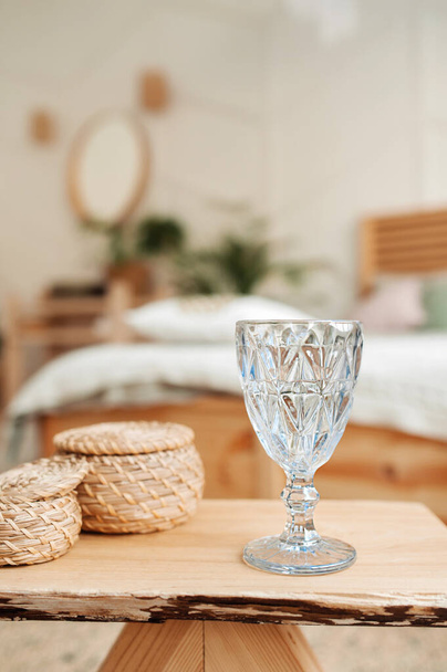 szklany kielich na drewnianym, ekologicznym stole z wiklinowymi koszykami na jasnym wnętrzu skandynawskiego stylu. zdrowy nawyk picia czystej wody rano, zawartość pionowa, wybiórcze skupienie - Zdjęcie, obraz