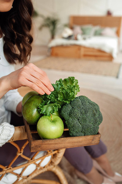 unrecognizable woman in white clothes holds in hands wooden eco storage box with green detox foods: apple, broccoli, lettuce, avocado. healthy diet food, vertical food content, selective focus - Foto, Imagem