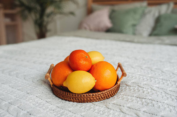 viele Zitrusfrüchte Orangen, Mandarinen und Zitronen auf hölzernem Weidenkorbtablett auf leichter Strickdecke auf gemütlichem Boho-Bett. gesunde Ernährung und Stärkung, selektiver Fokus - Foto, Bild