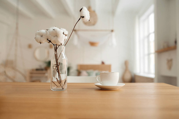 weiße Porzellantasse und Bouquet aus natürlicher Baumwolle in Glasvase auf hölzerner Arbeitsplatte im hellen Inneren der Wohnung im Boho-Stil. Selektiver Fokus - Foto, Bild