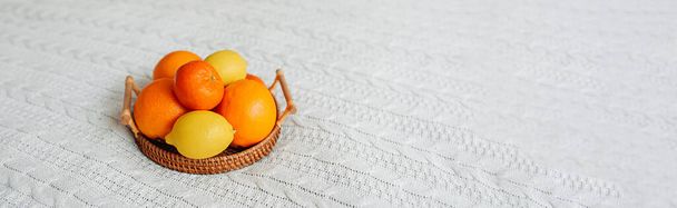 Großformat Banner viele Zitrusfrüchte Orangen, Mandarinen und Zitronen auf hölzernen Weidenablage auf leicht gestrickte Decke auf gemütlichen Boho-Bett. gesunde Ernährung und Stärkung, selektiver Fokus - Foto, Bild