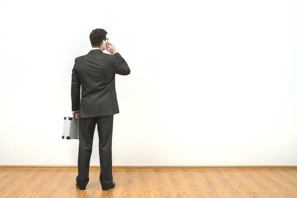 Ο επιχειρηματίας με το χαρτοφύλακα στο φόντο του λευκού τοίχου. - Φωτογραφία, εικόνα