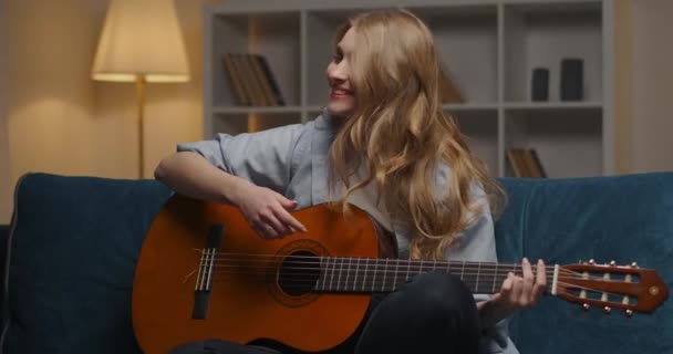Χαρούμενη νεαρή κοπέλα με κιθάρα διασκεδάζει στο σπίτι το βράδυ, κάθεται μόνη της στο άνετο διαμέρισμα και απολαμβάνοντας μουσική - Πλάνα, βίντεο