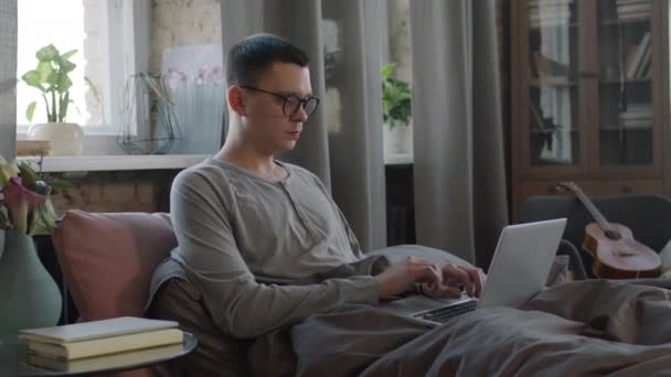 Plan moyen de pigiste dans les vêtements à la maison tapant sur ordinateur portable alors qu'il était assis au lit dans un appartement loft lumineux le matin - Séquence, vidéo