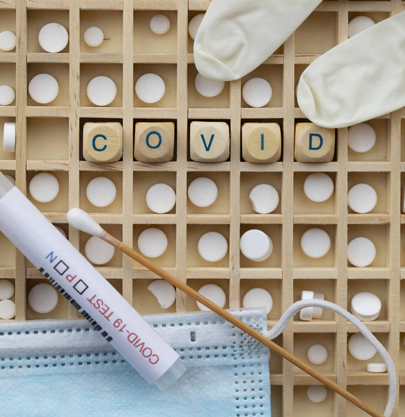 Слово COVID состоит из деревянной буквы. Маска и мазок из носа на заднем плане - Фото, изображение