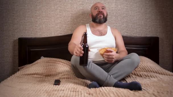un uomo barbuto ben nutrito di aspetto europeo siede a casa sul letto in abiti casual, beve birra da una bottiglia e mangia patatine fritte. c - Filmati, video