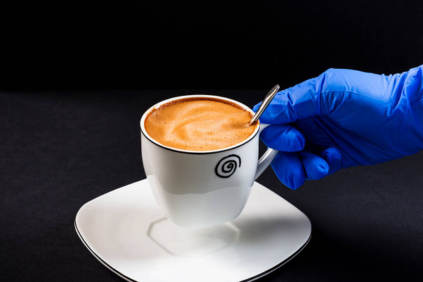 Person's hand with latex glove raising a cup of latte.This is a horizontal format photo taken against a black background under artificial lighting in a studio. - Photo, Image